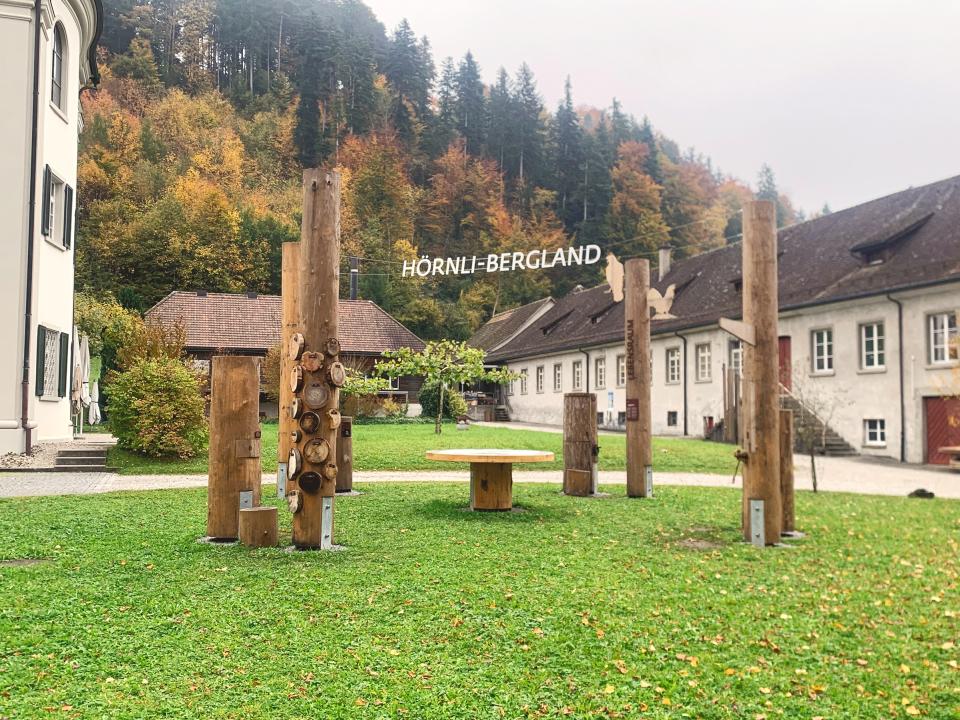 Baumstämme auf einer Wiese auf dem Klostergelände, mit Drähten verbunden, an denen Buchstaben das Wort Hörnli-Bergland ergeben