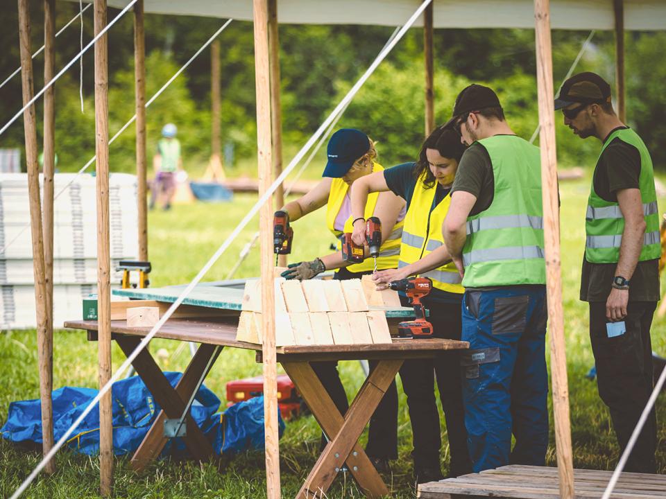 Vier junge Menschen helfen beim Aufbau des Jublasuriums; sie haben Bohrmaschinen in der Hand und Leuchtwesten
