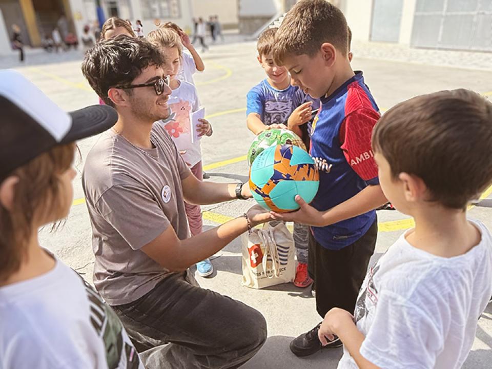 Junger Mann kniet vor Kindern, hält einem Jungen einen Ball hin