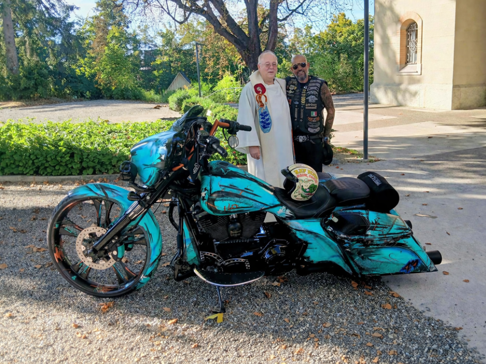 Motorrad in ozeanblauer Farbe, dahinter ein Priester und ein tätowierter Biker mit Sonnenbrille