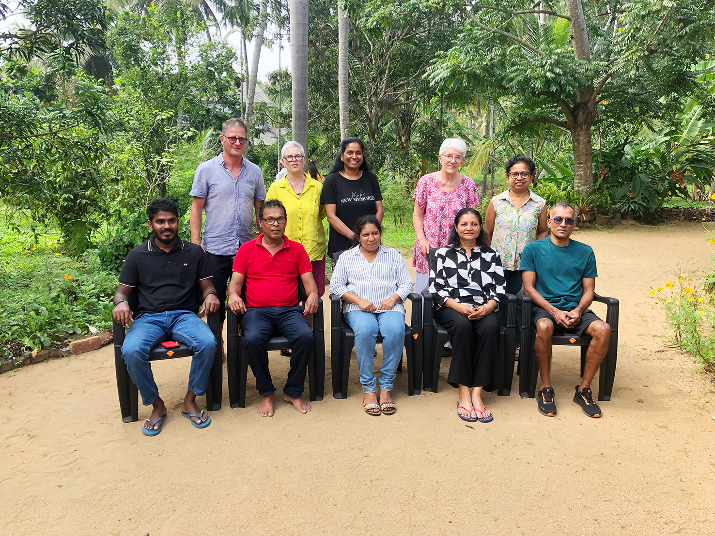 fünf sitzende Personen in Sri Lanka sind das Helferteam des Vereins Lankawelamai im Distrikt Kurunegala, dahinter stehend der fünfköpfige Schweizer Besuch des Vereins
